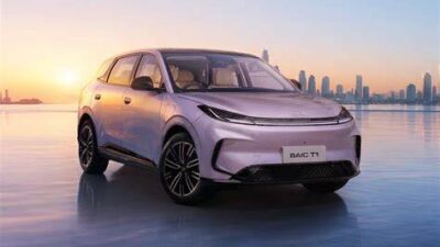 White futuristic SUV photographed from the front-left, parked on a reflective icy surface with a sunset and city skyline in the background.