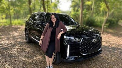 Woman in a maroon cardigan standing beside a black SUV in a wooded parking area.