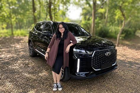 Woman in a maroon cardigan standing beside a black SUV in a wooded parking area.