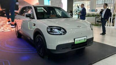 White compact Chery car on a showroom platform, front angled to the camera with blue lighting around the display and people in the background inspecting the vehicle.
