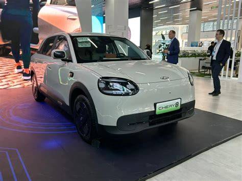 White compact Chery car on a showroom platform, front angled to the camera with blue lighting around the display and people in the background inspecting the vehicle.