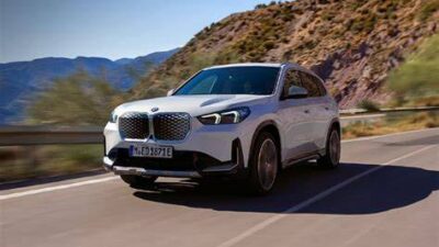 White BMW SUV driving along a winding mountain road with rocky slopes and clear blue sky in the background.