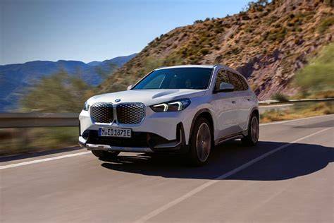 White BMW SUV driving along a winding mountain road with rocky slopes and clear blue sky in the background.